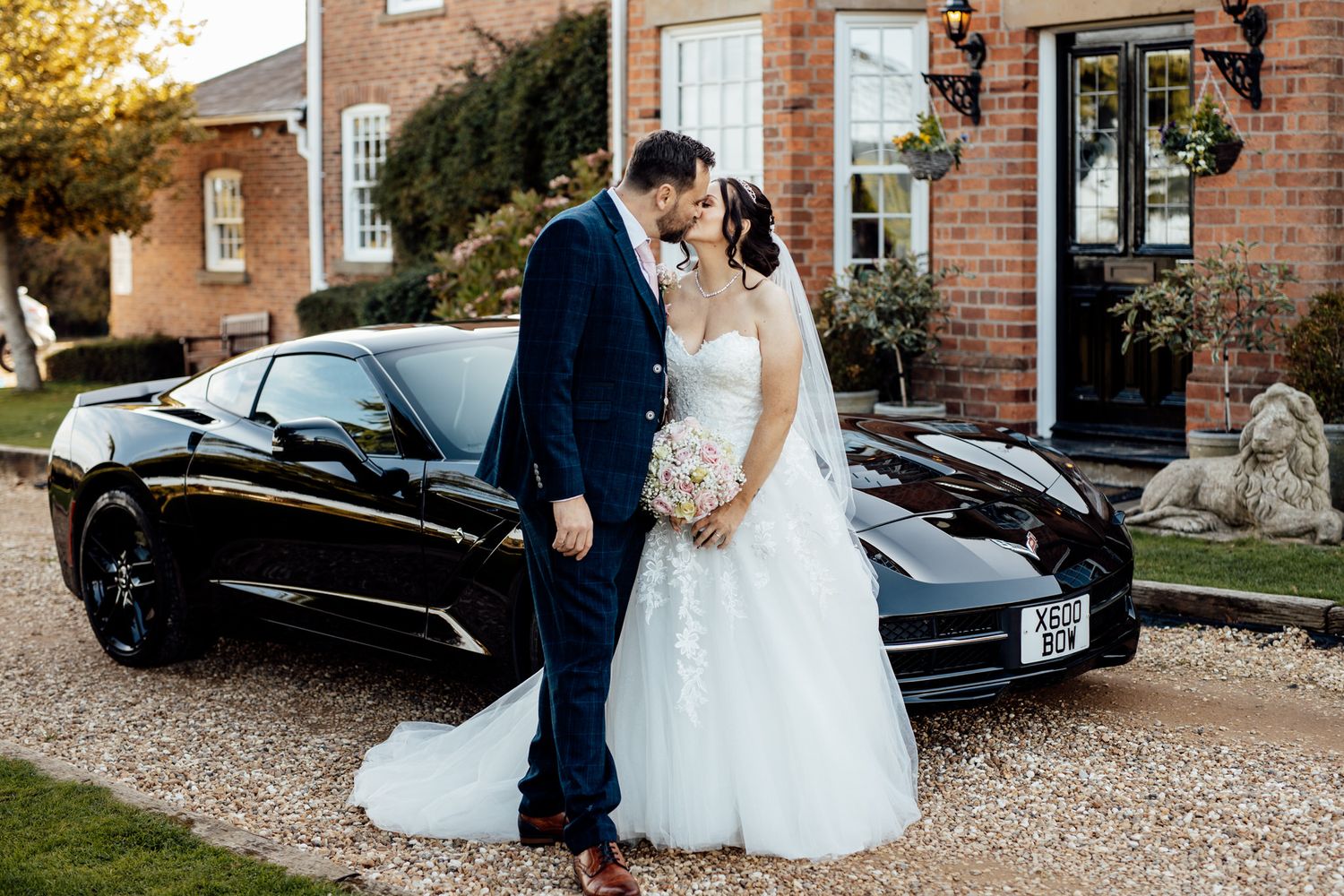 Elegant wedding couple poses by vintage car outside brick venue at golden hour.