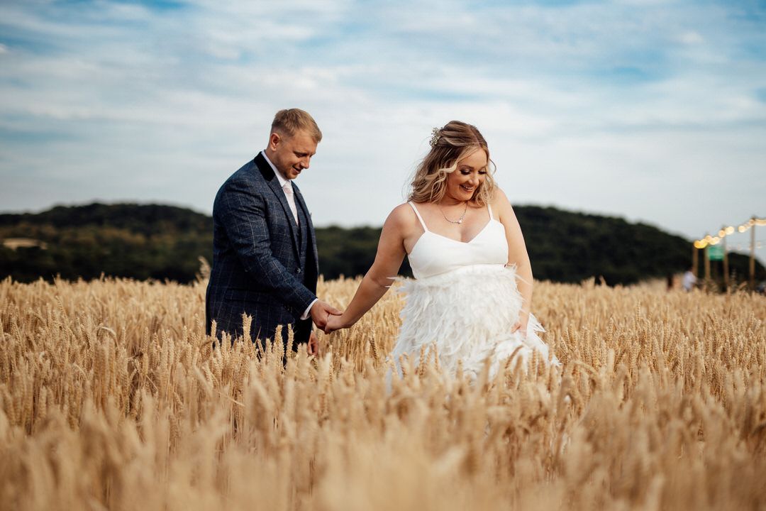 Newlyweds walk hand in hand through a golden wheat field under blue skies on their wedding day.