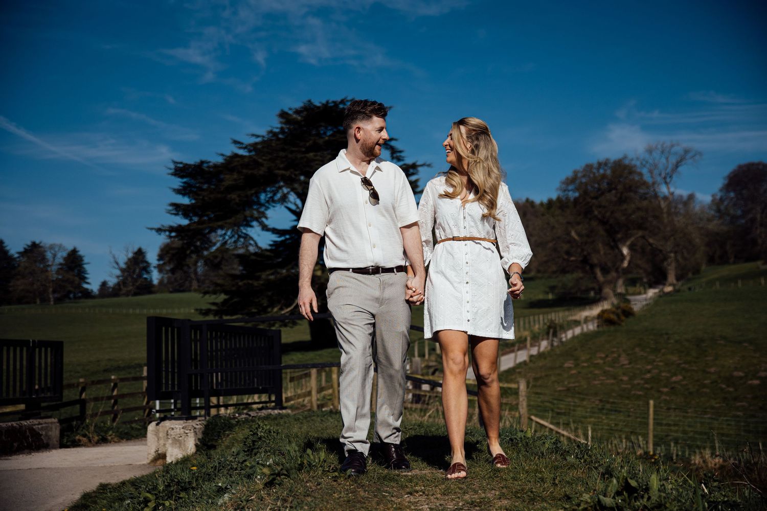 A couple walking hand in hand on a path under bright blue skies and rolling hills.