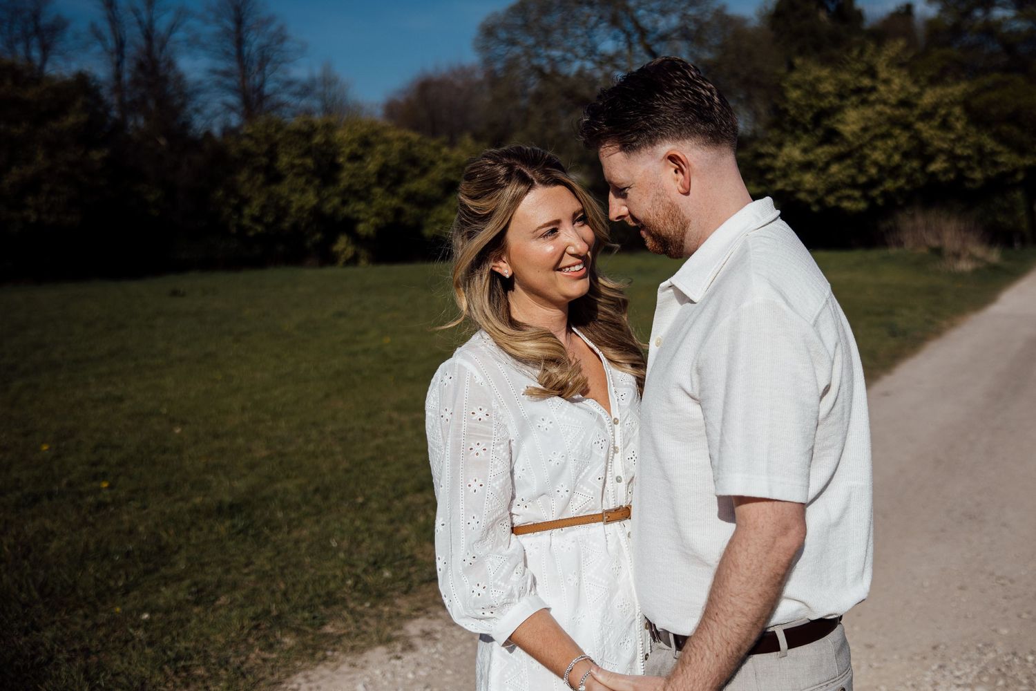Couple in coordinated white outfits share intimate moment on sunny park path.