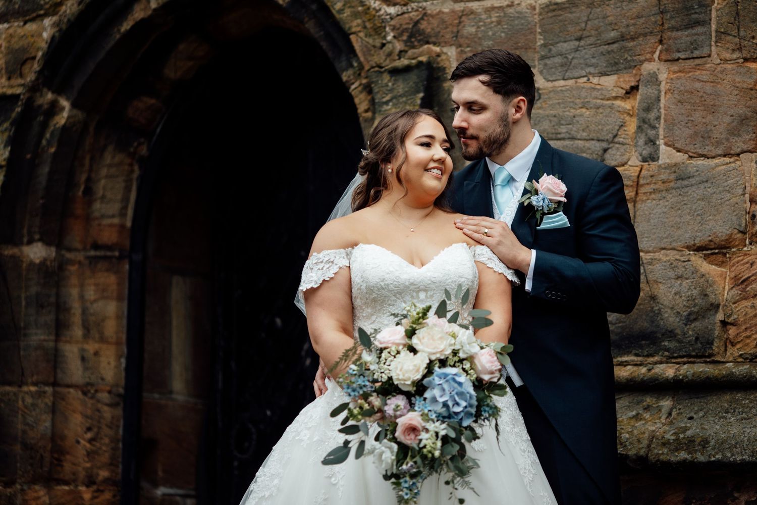Newlyweds pose together against stone church wall with blue and white floral bouquet.