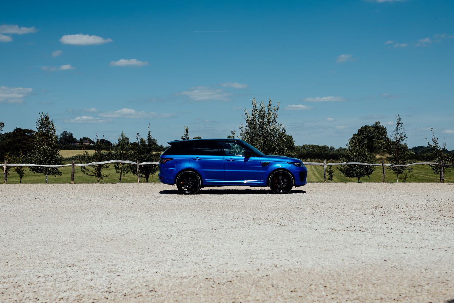Blue sports car parked on white gravel surface against scenic rural landscape on sunny day.