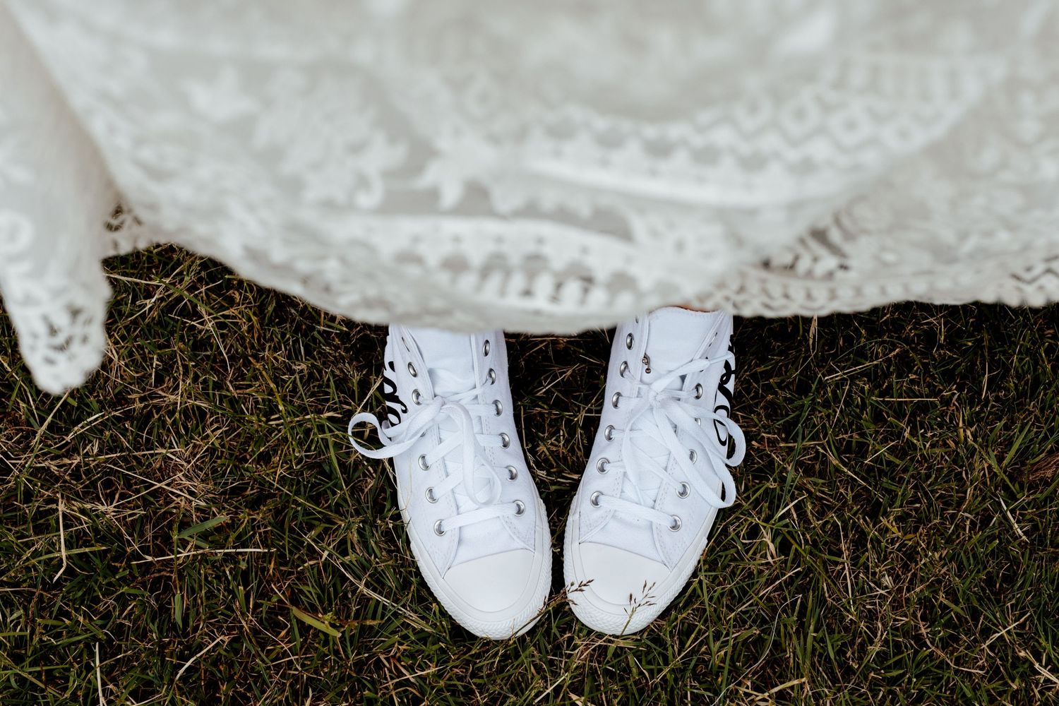 White lace-up sneakers peeking out from under wedding dress on grass.