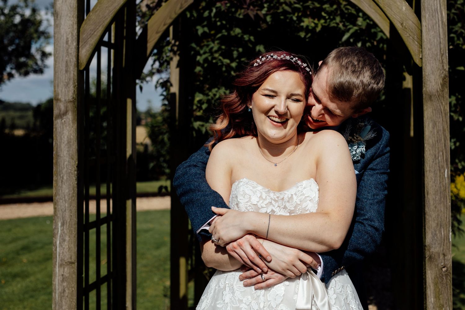 Romantic moment captured under wooden garden arch during summer wedding.
