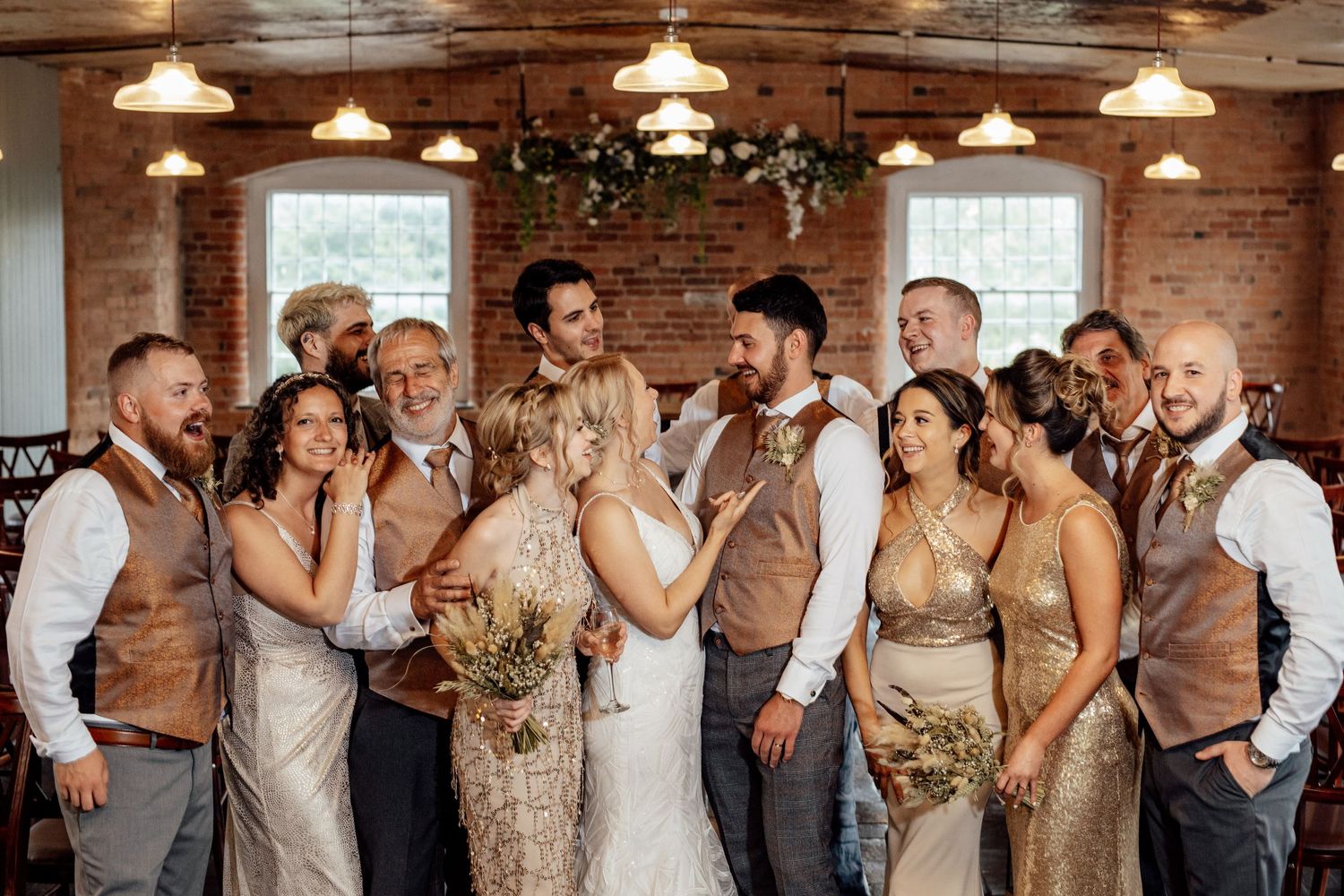Wedding party in neutral tones and vests posing together in rustic venue with pendant lights.