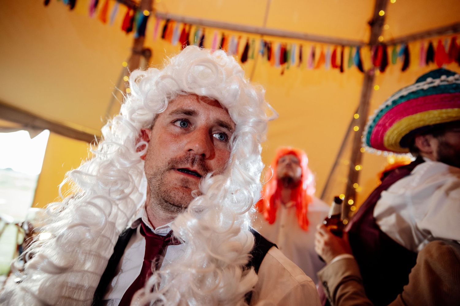 Party guest wearing a white wig and tie at a festive celebration with colorful decorations.
