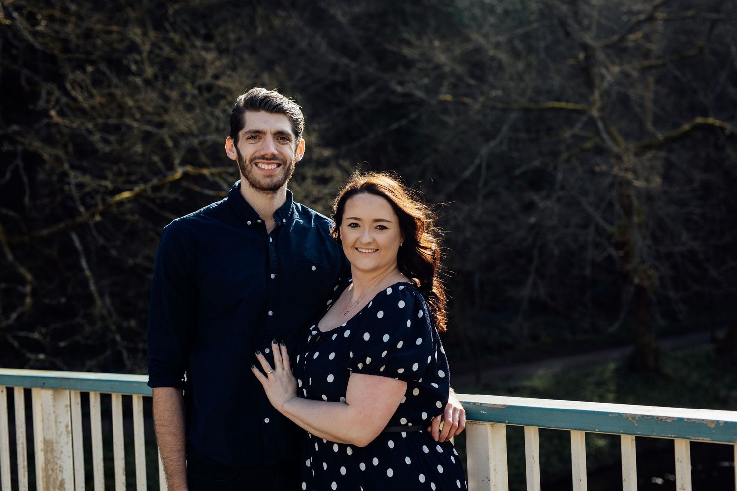 A couple poses on a bridge with scenic mountain views in the background.