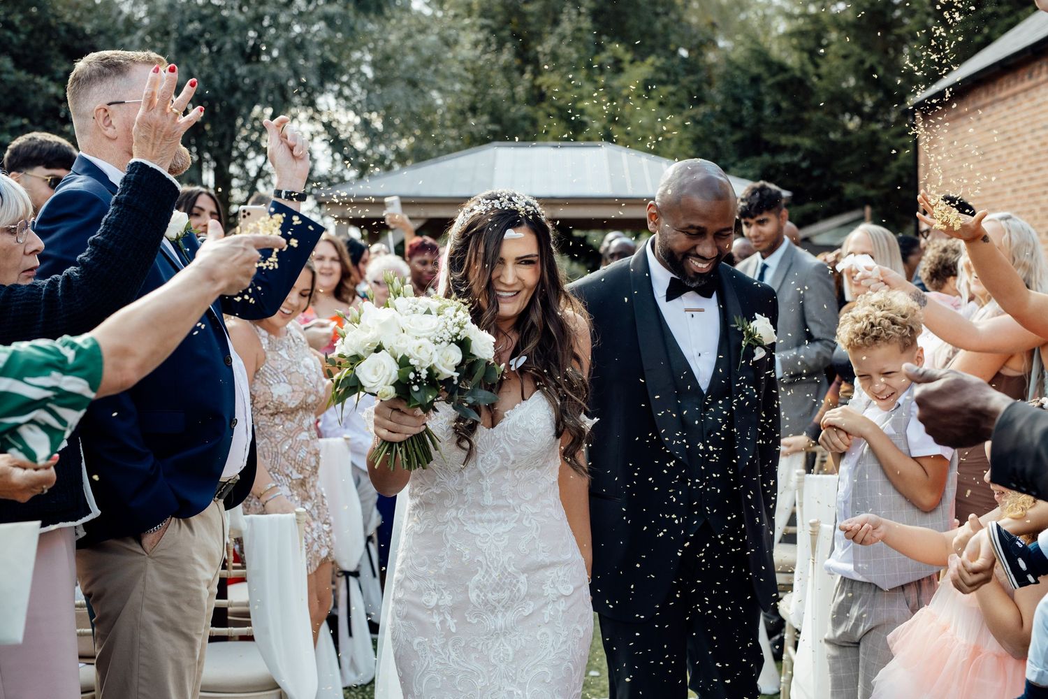 Wedding guests celebrate and throw confetti as the newlyweds walk down the aisle together.