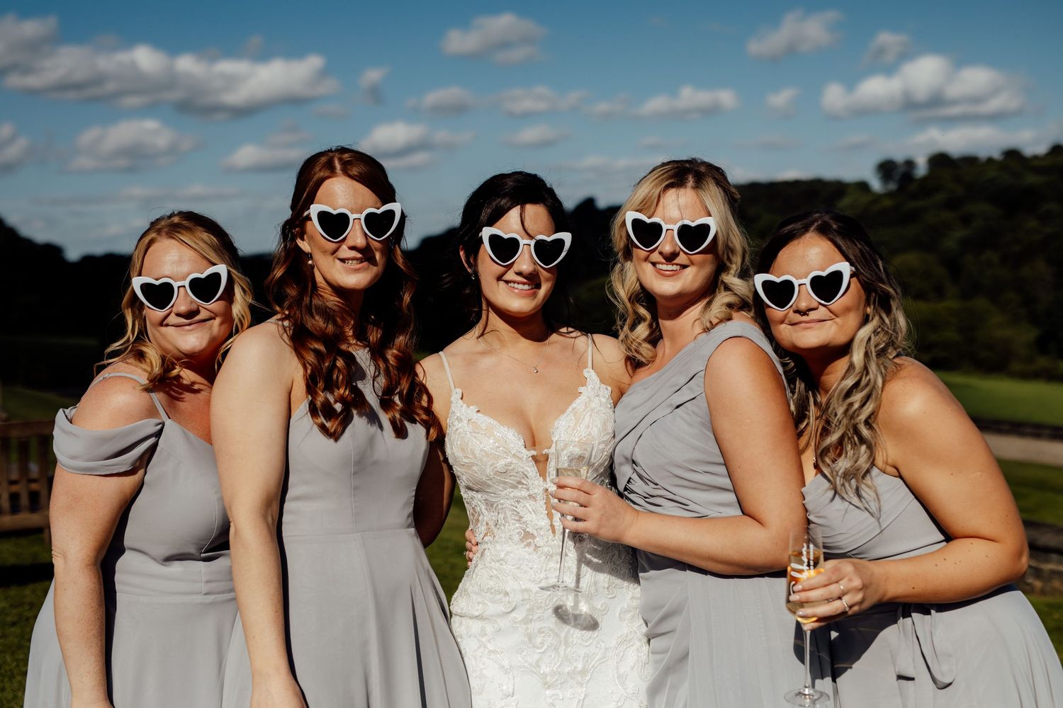 Bridesmaids in matching gray dresses wearing sunglasses pose outdoors on a sunny day.