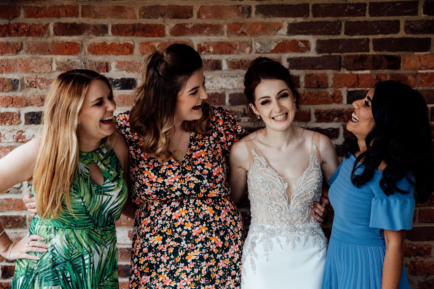 Wedding party in floral and blue dresses pose together against rustic brick wall backdrop.