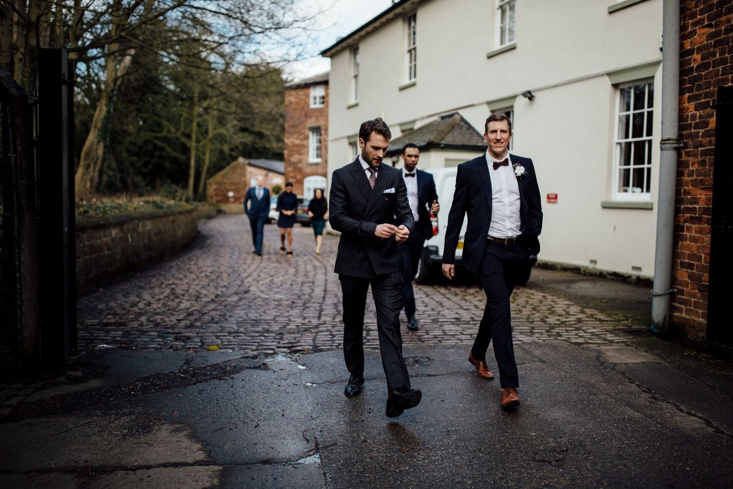 Wedding guests in formal attire walk along a brick pathway outside a historic building.