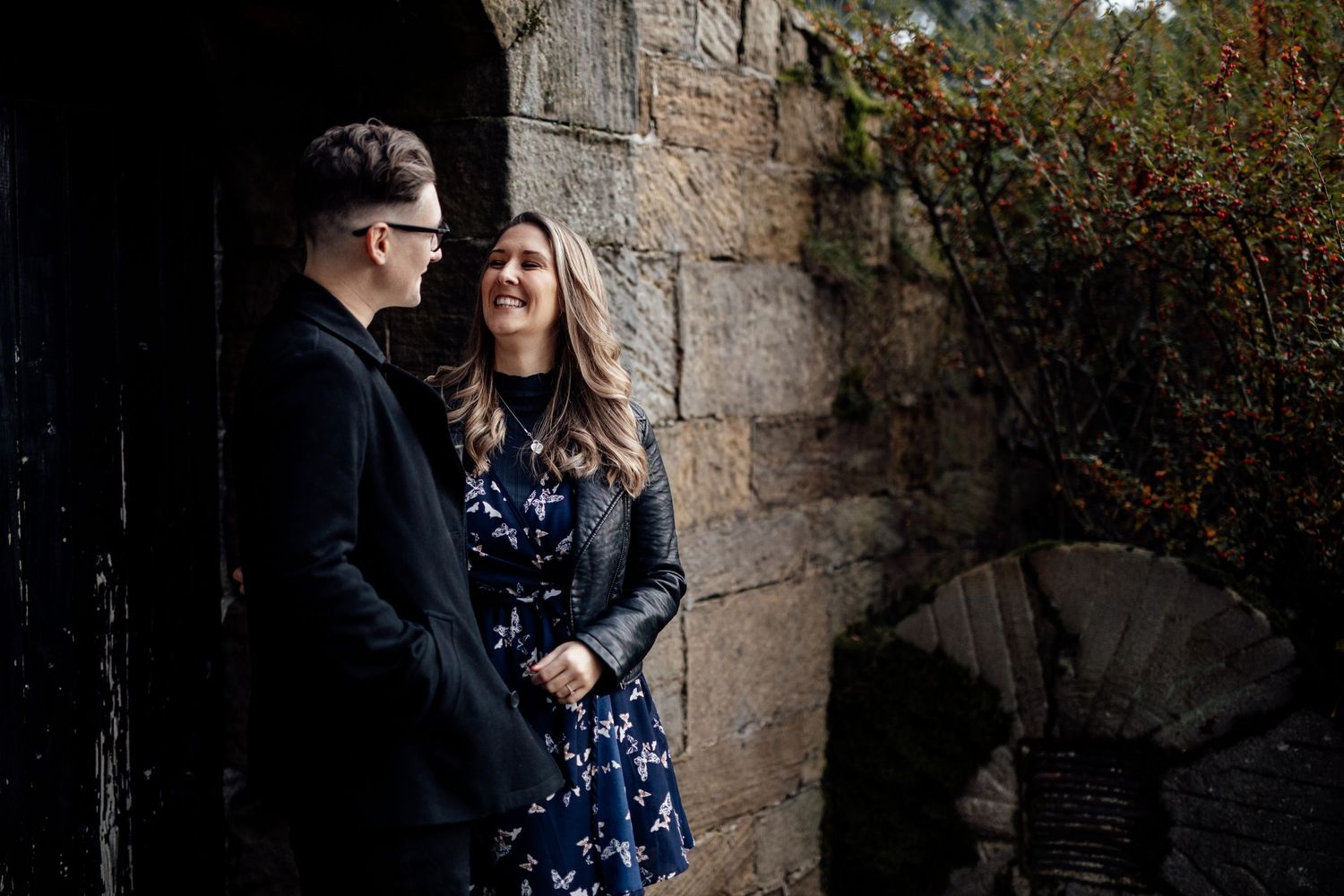 A couple poses against a stone wall with greenery during an autumn engagement shoot.