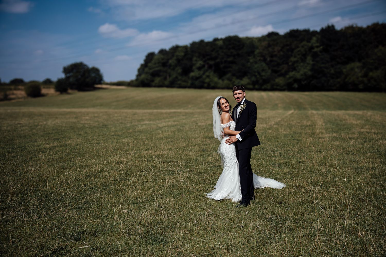 A wedding couple poses in a green field with trees in the background on their special day.