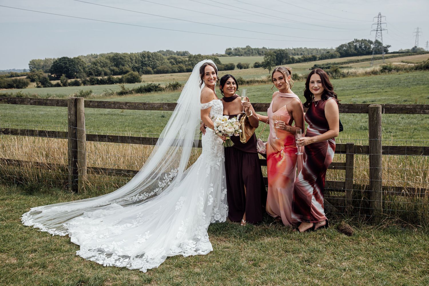 A bridal party in burgundy dresses poses with the bride in front of a countryside fence.
