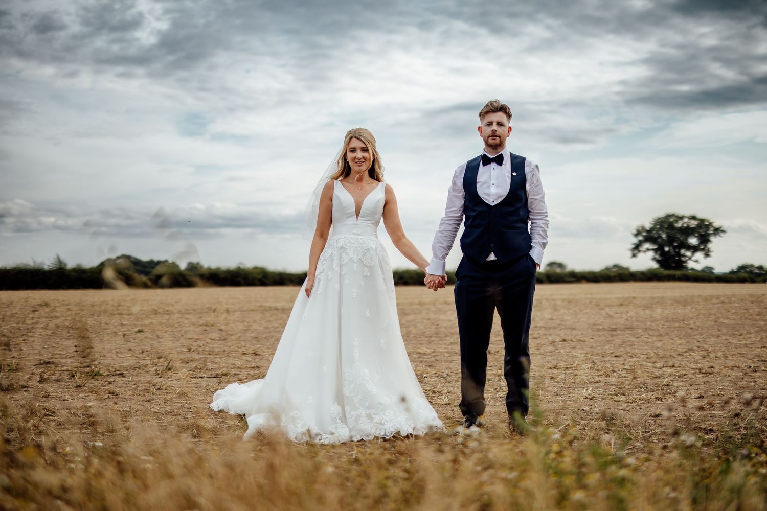 Newlyweds walk hand in hand through an open field under cloudy skies.