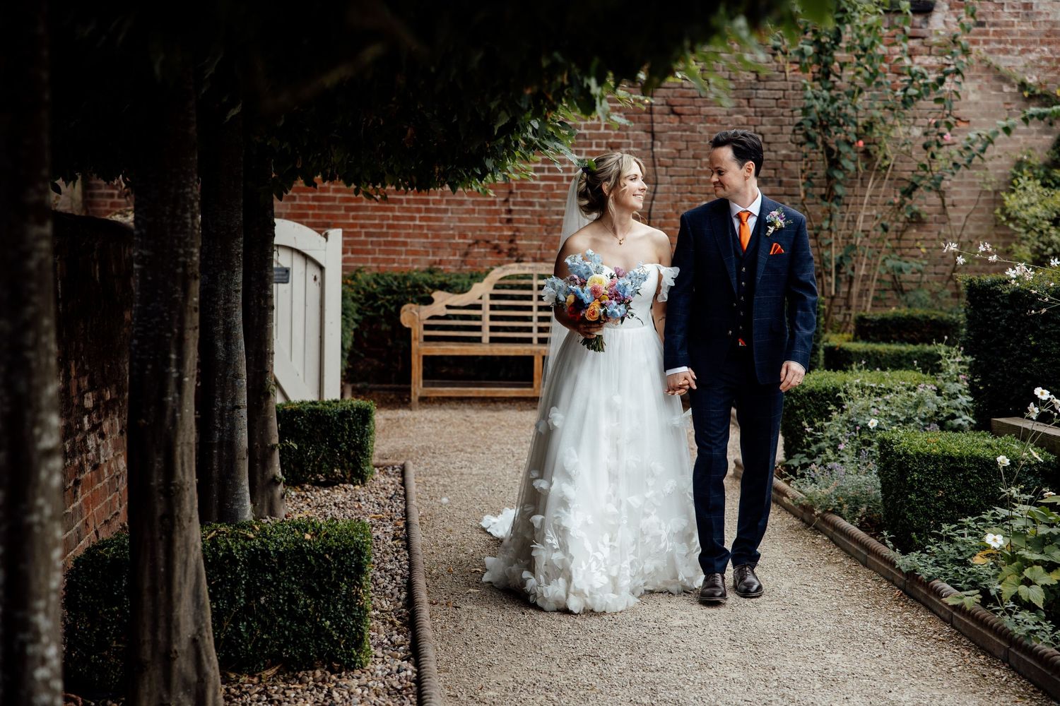 Wedding party walking through a garden path lined with brick walls and trees.