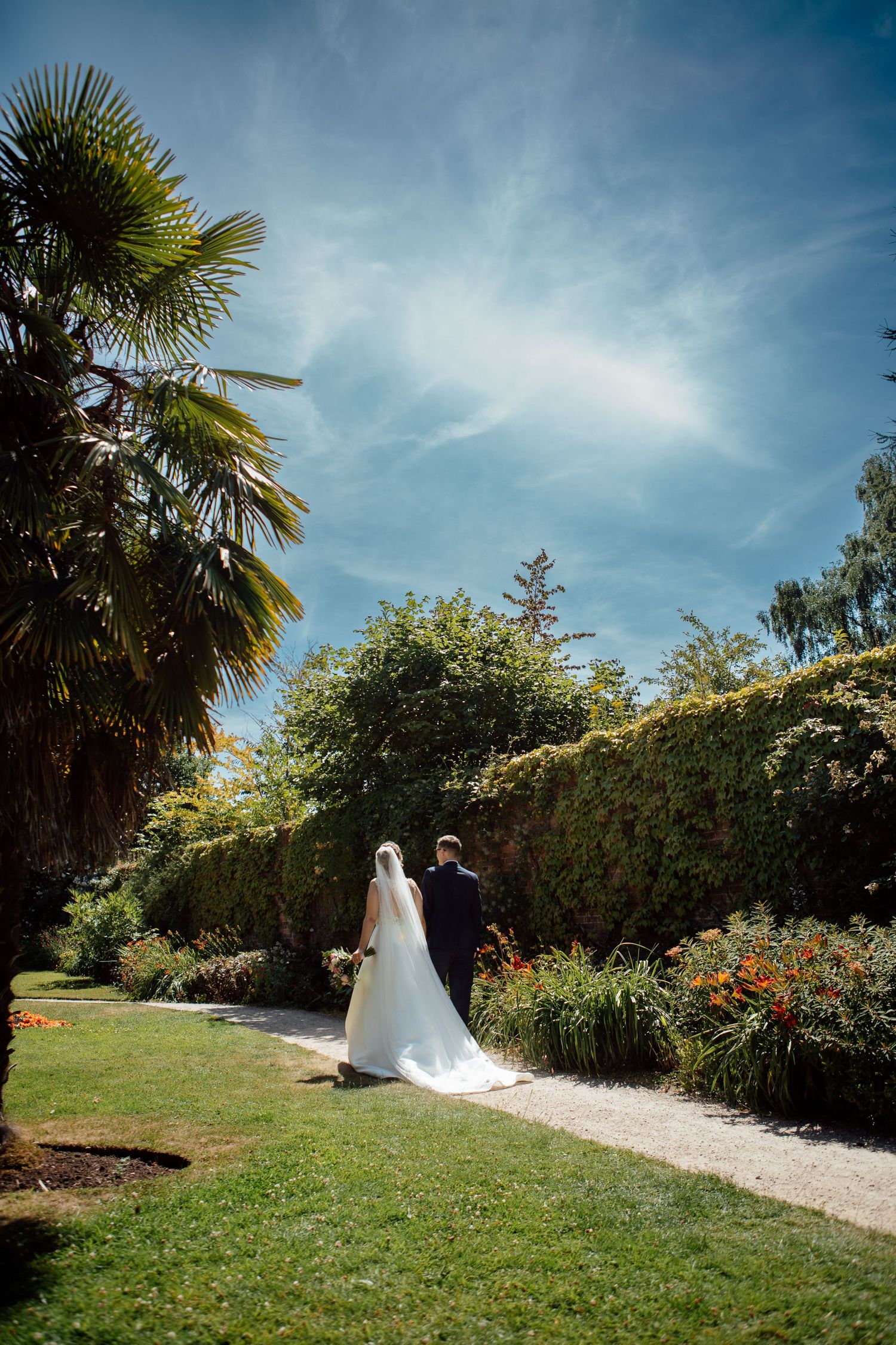 Wedding couple walks along a garden path with palm trees and blooming flowers on a bright summer day.