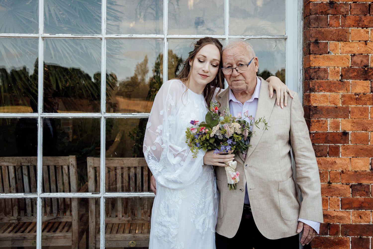 Wedding couple poses against rustic brick wall with white windows in elegant countryside venue setting.