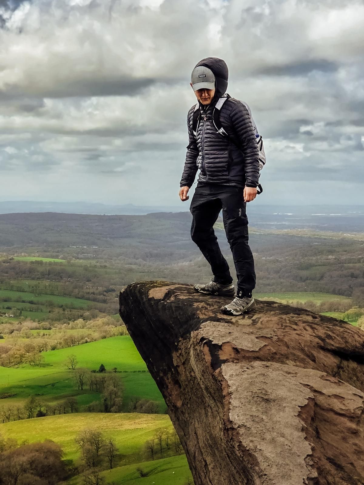 Hiker standing on rocky cliff edge overlooking lush green valley and rolling hills under dramatic cloudy sky.