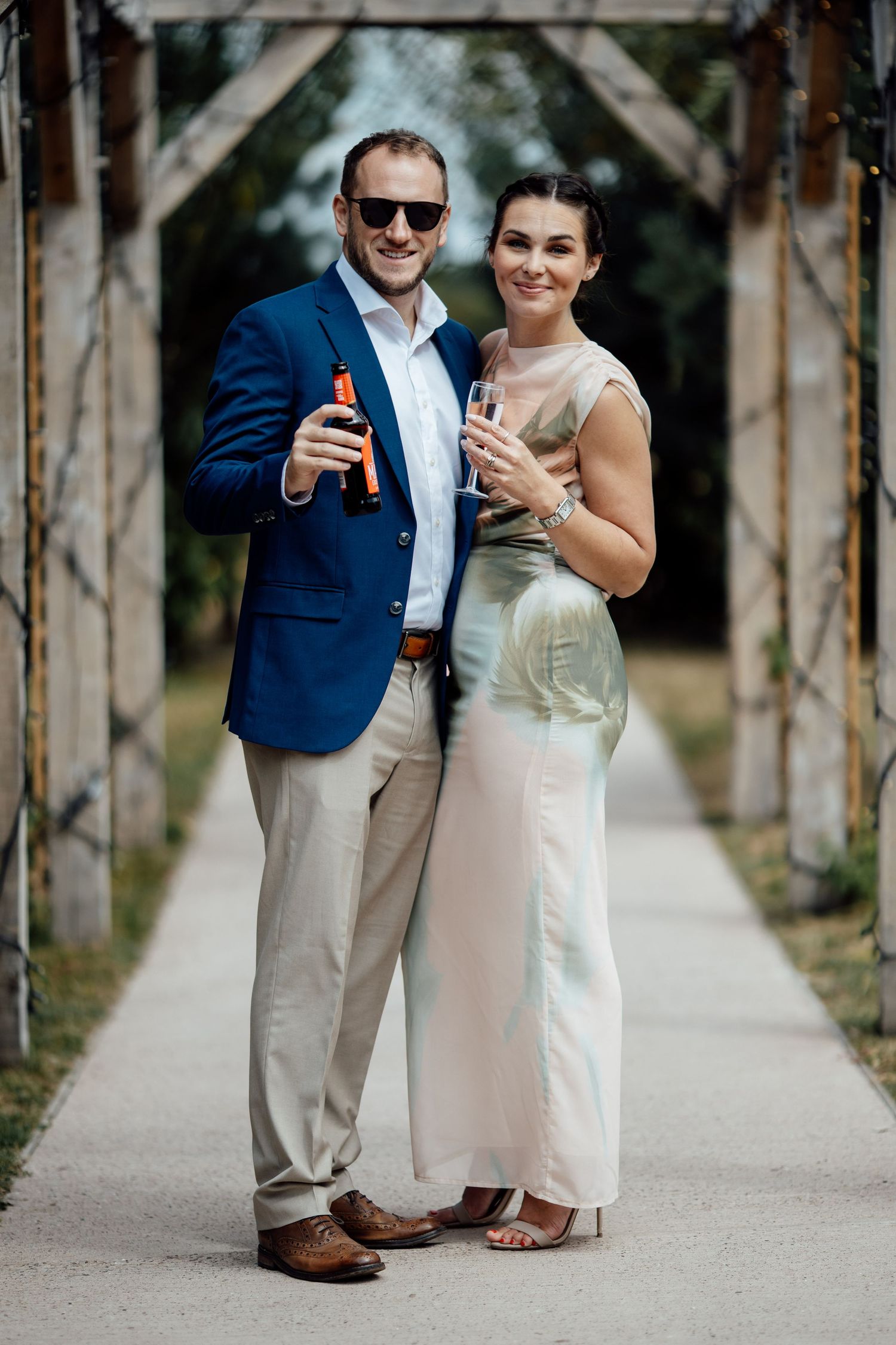 Couple in formal wear toasting with champagne bottle under wooden arch outdoors.