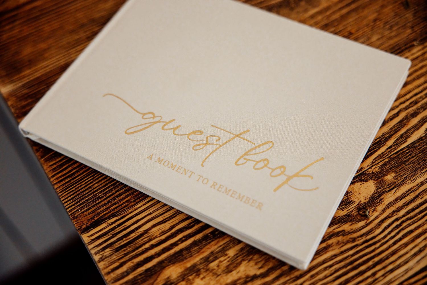 Elegant white wedding guest book with gold script reading guest book a moment to remember on wooden table.