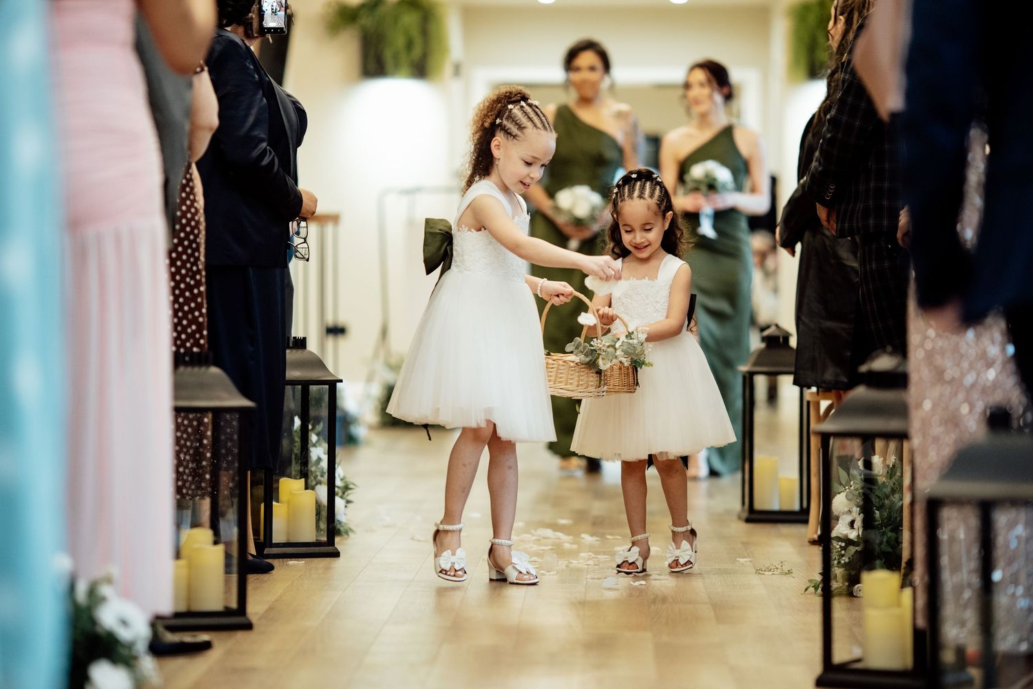 Two flower girls in white dresses walk down the wedding aisle carrying a floral basket.