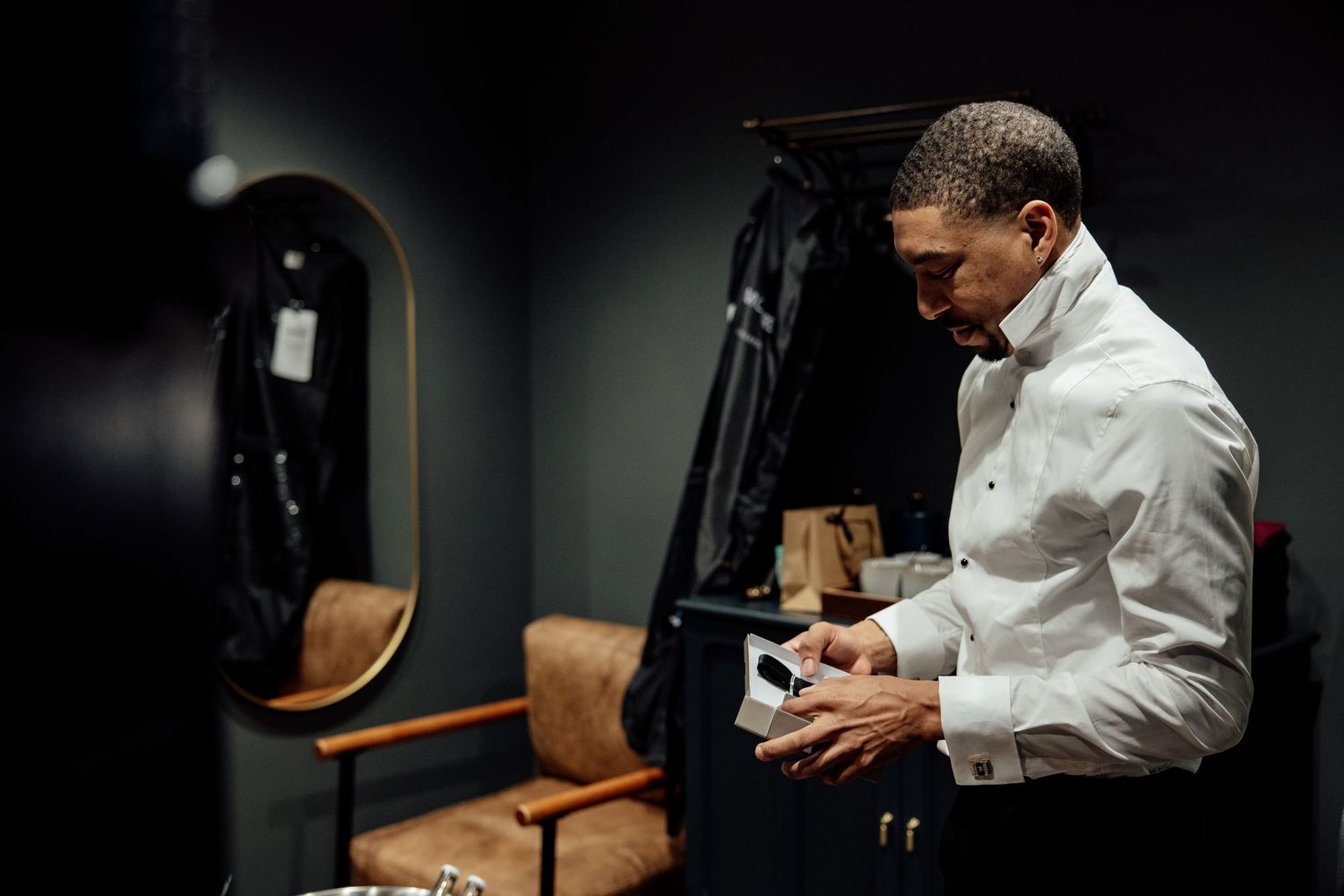 A groom in a white dress shirt adjusts his cufflinks in a dimly lit dressing room before his wedding.
