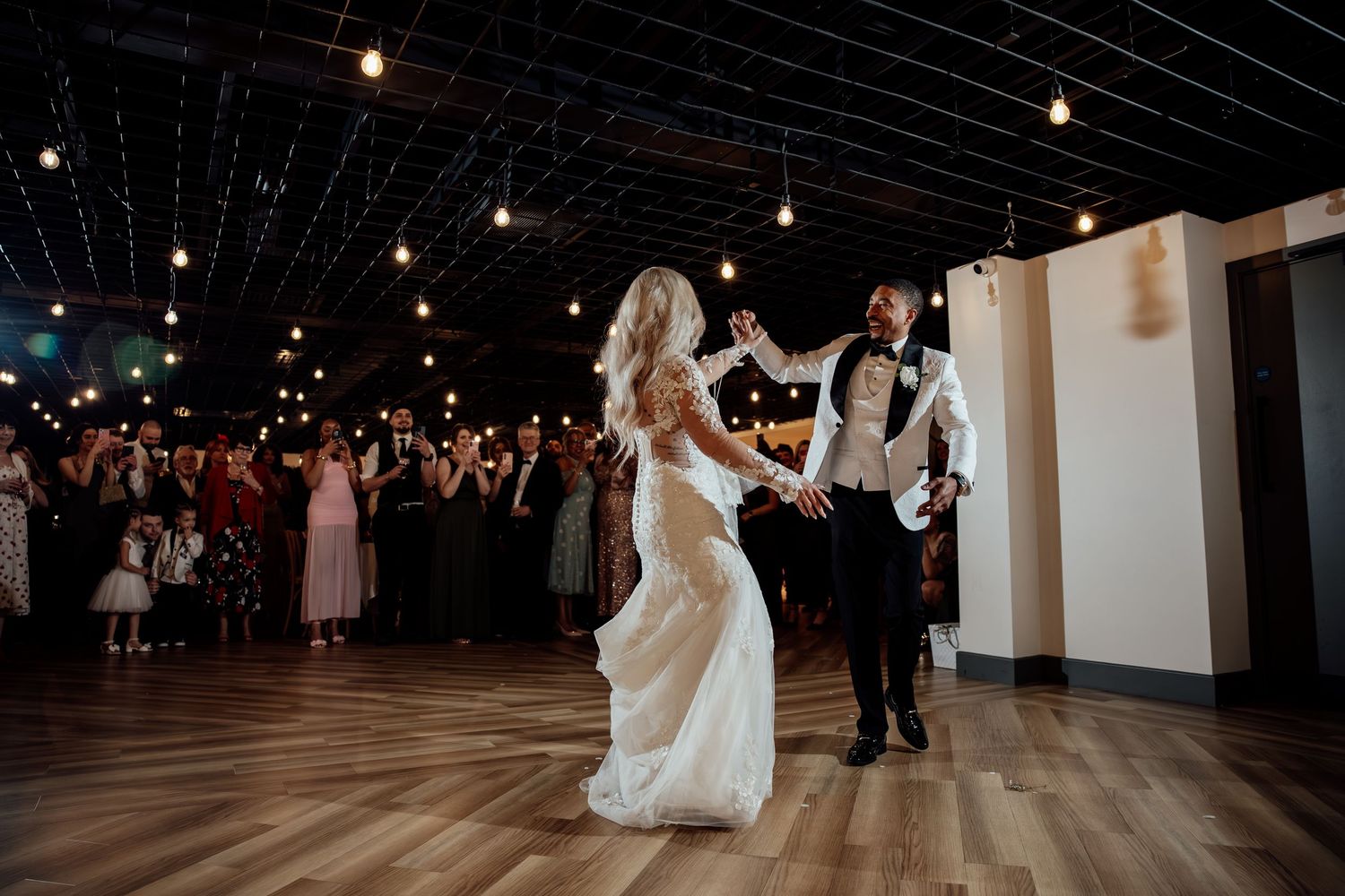 Bride and groom share their first dance surrounded by cheering guests in a warmly lit wedding venue.
