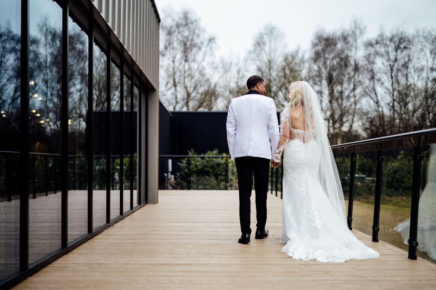 Bride in backless wedding gown and groom in white tuxedo walking on modern venue deck.