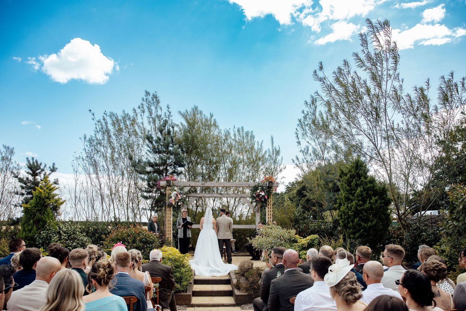 Outdoor wedding ceremony with bride and groom at floral arch, guests seated on sunny day with blue sky and trees.