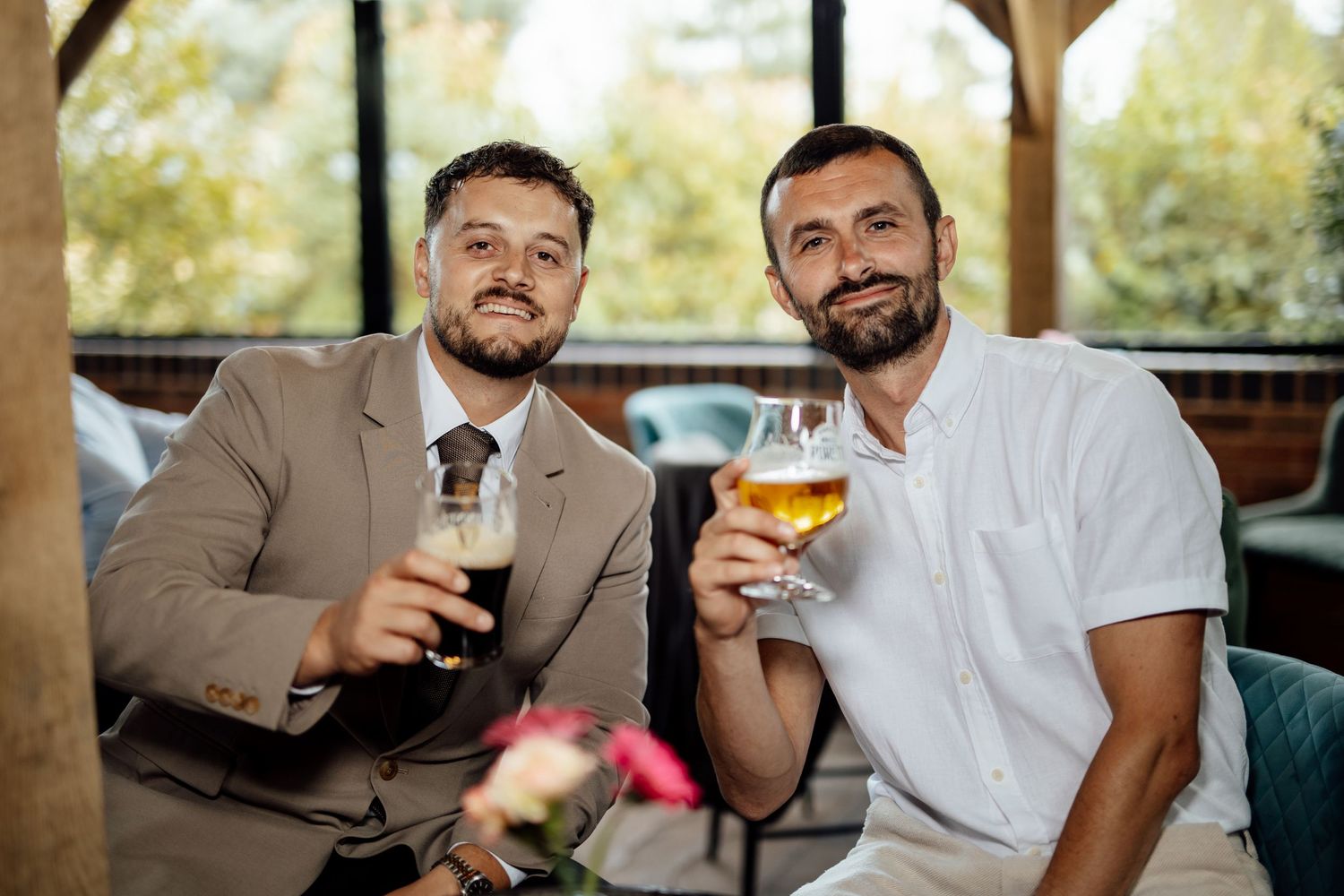 Two well-dressed men smiling and toasting with beer glasses at an outdoor wedding reception.