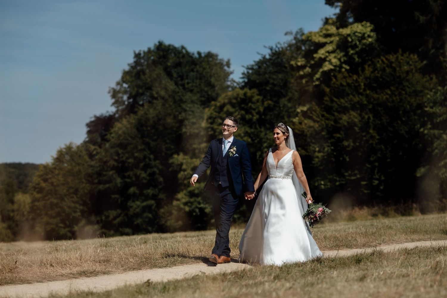 A bride and groom walk together through a scenic meadow on their wedding day during golden hour.