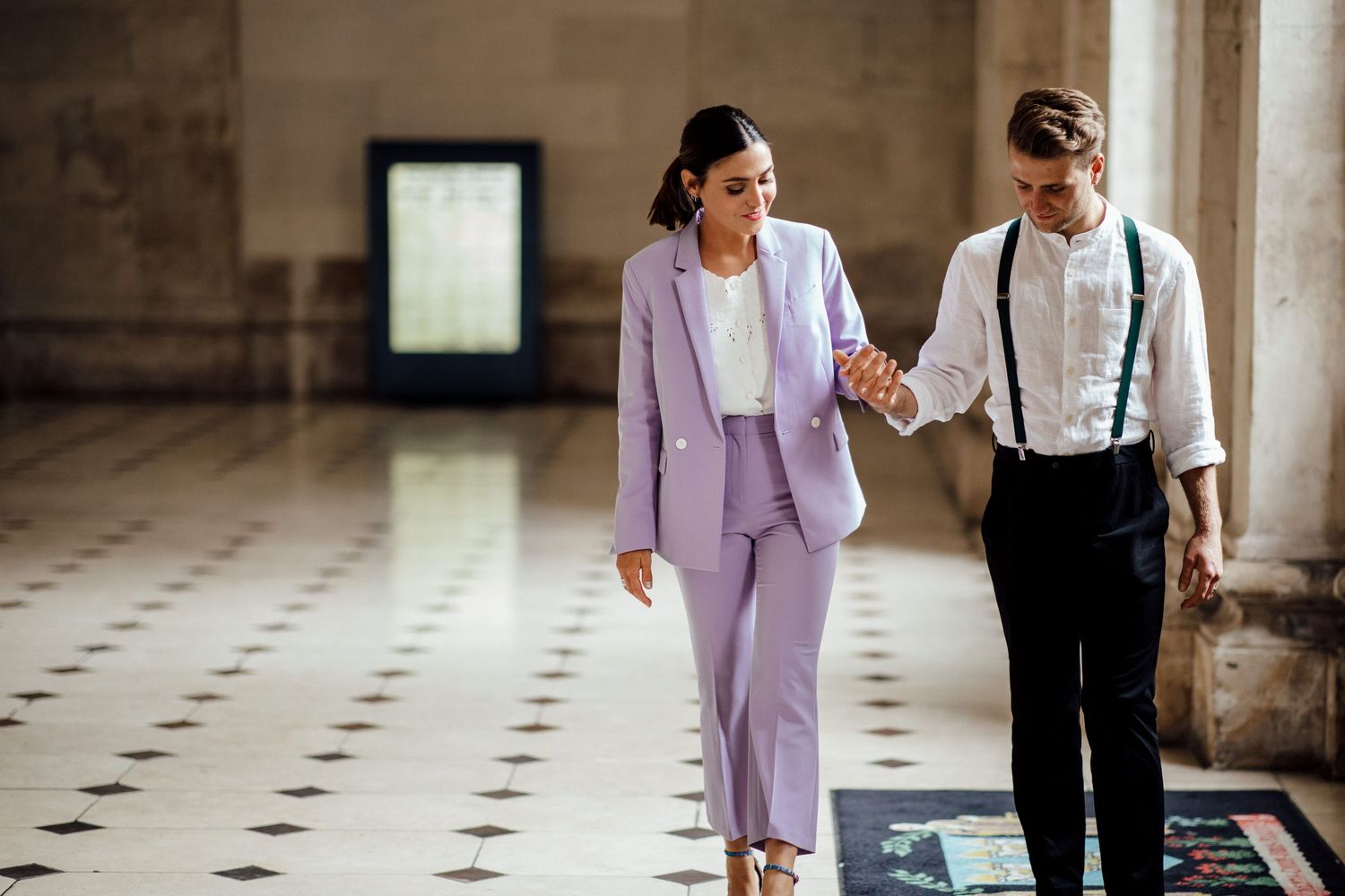 Couple walking hand in hand through a grand hallway with tiled floors.