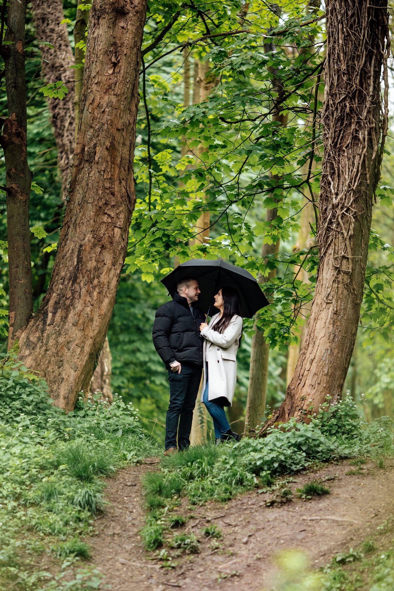 A couple shares a romantic moment under a black umbrella among tall trees.