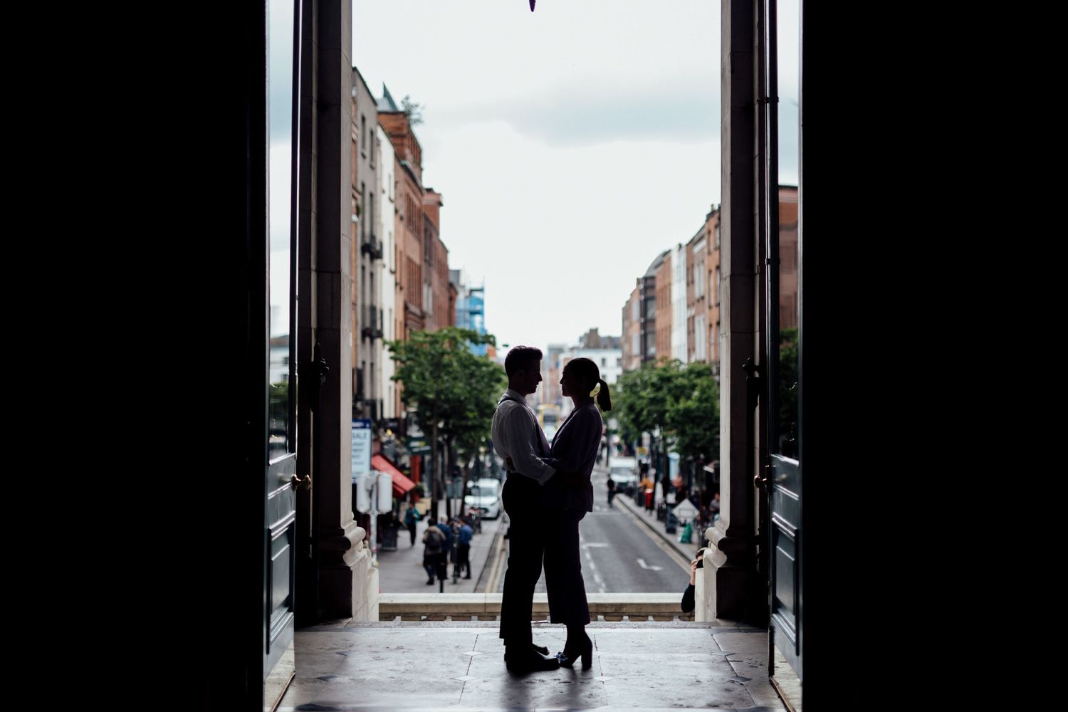 Silhouetted couple stands in a doorway overlooking a European city street lined with buildings.