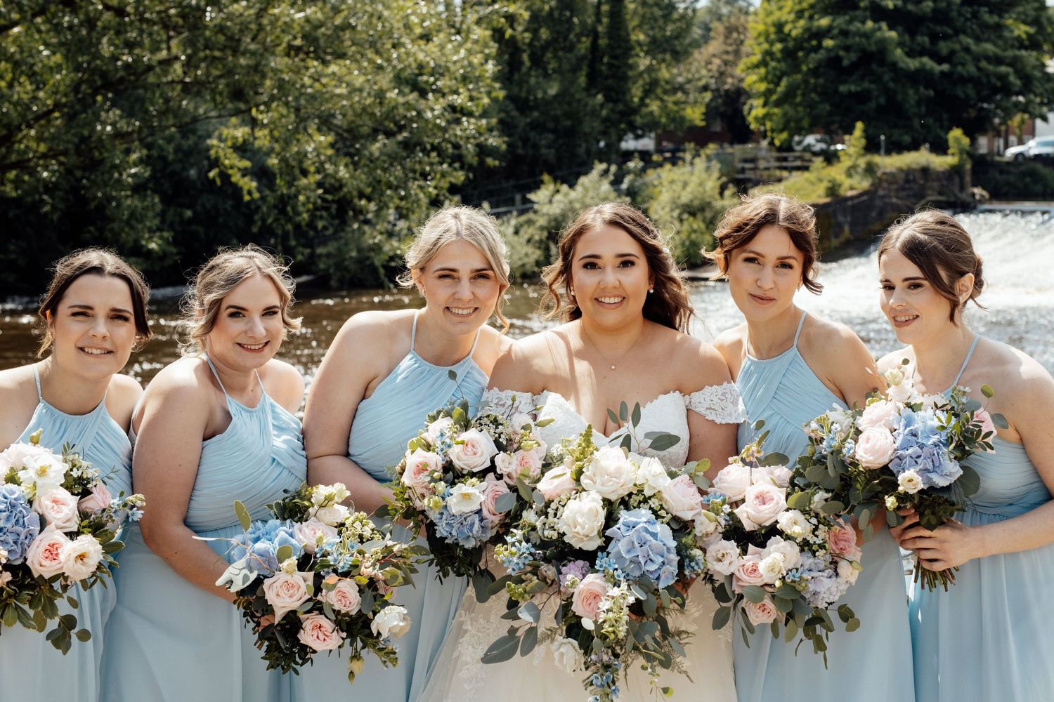 Bridal party in powder blue dresses holds lush floral bouquets in sunny outdoor setting.