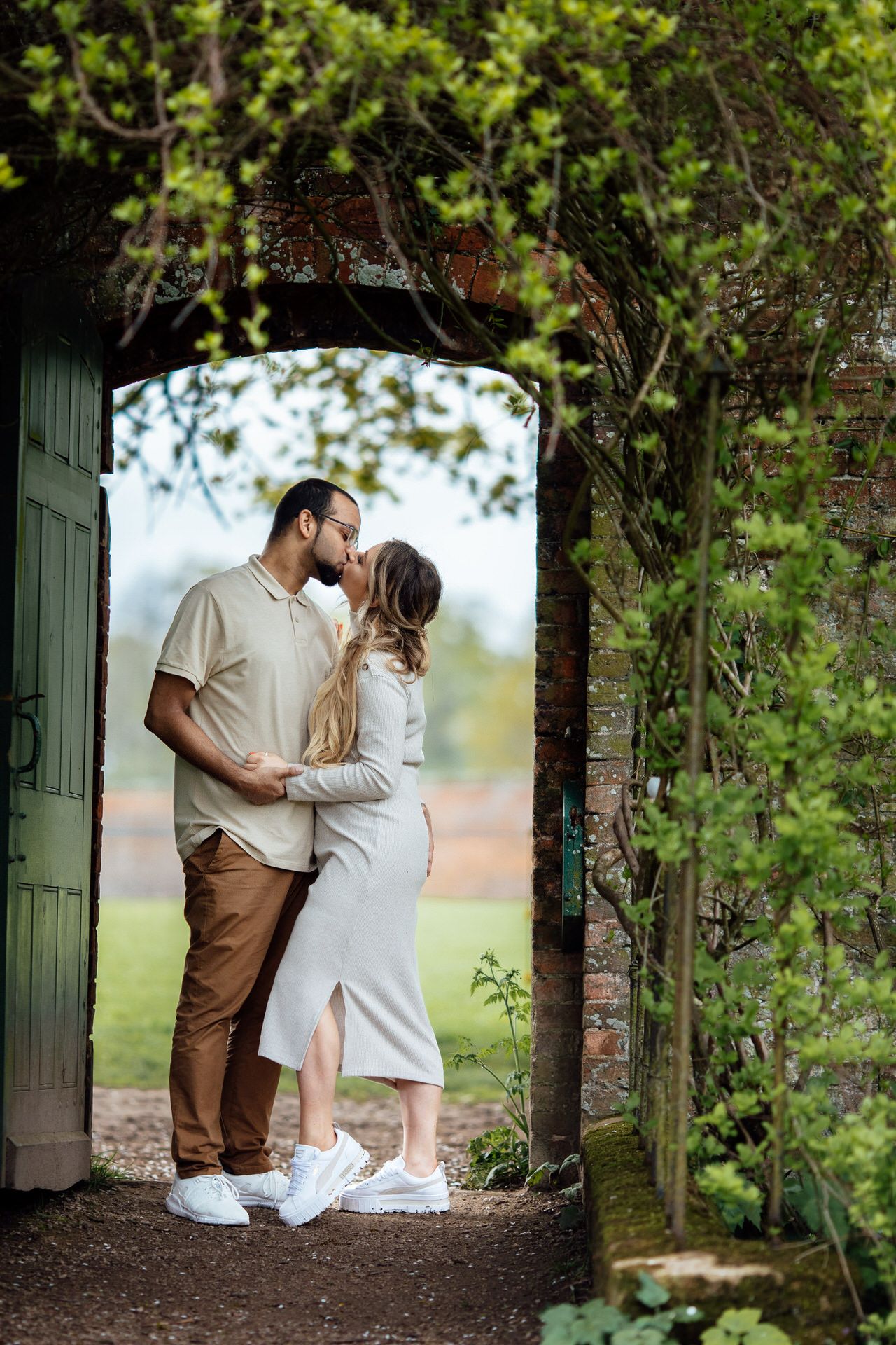 Romantic garden photoshoot with couple framed by lush green archway and wooden doors.