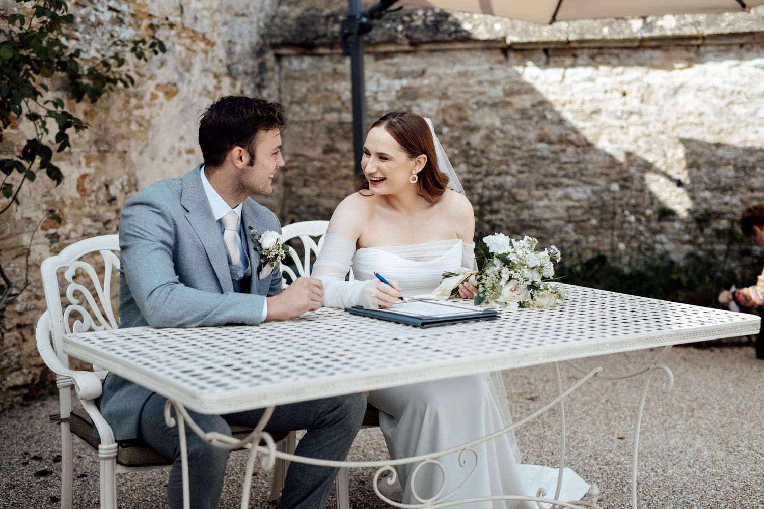Couple seated at white metal table during outdoor wedding celebration.