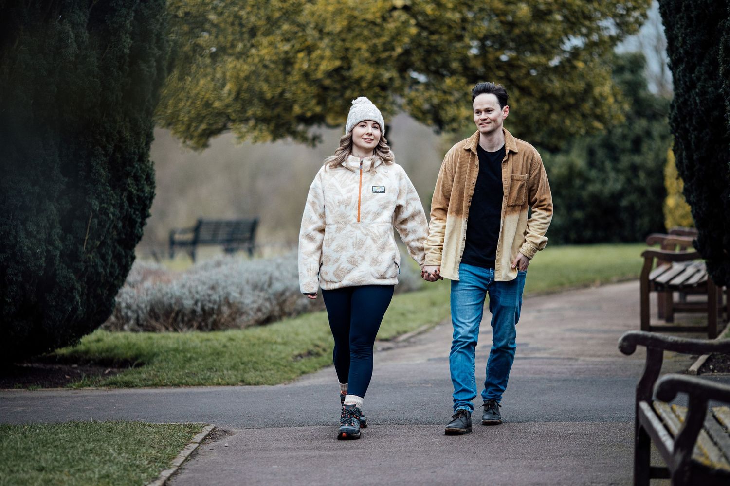 Couple strolling through a park pathway with benches and trees in the background.
