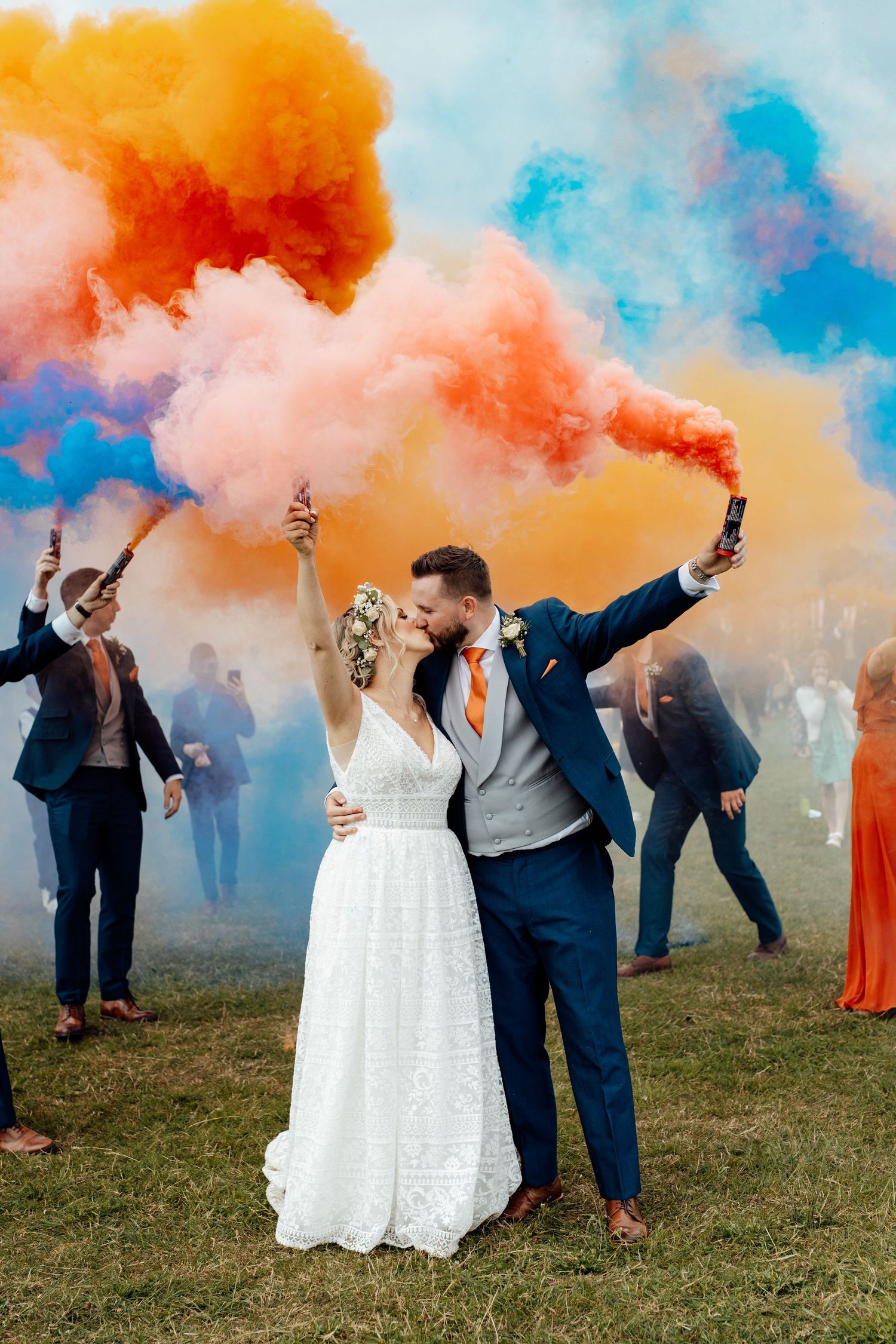 A couple embraces amid vibrant orange and blue smoke bombs during an outdoor celebration.
