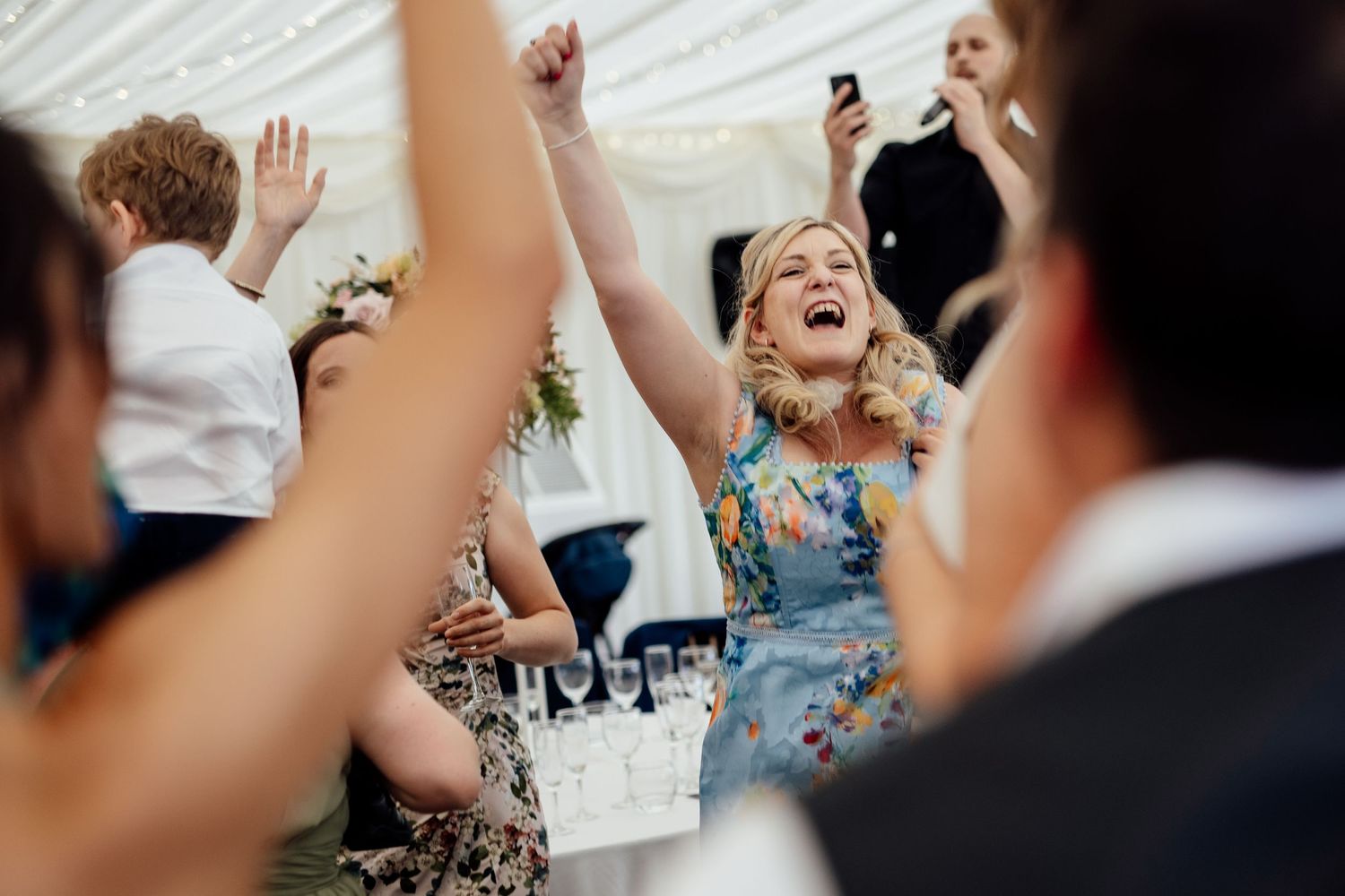 Wedding guests celebrate with raised arms and joyful expressions inside a white tent reception.