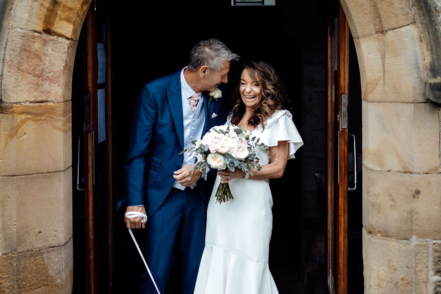 Couple exits stone church holding bouquet and walking stick after their ceremony.