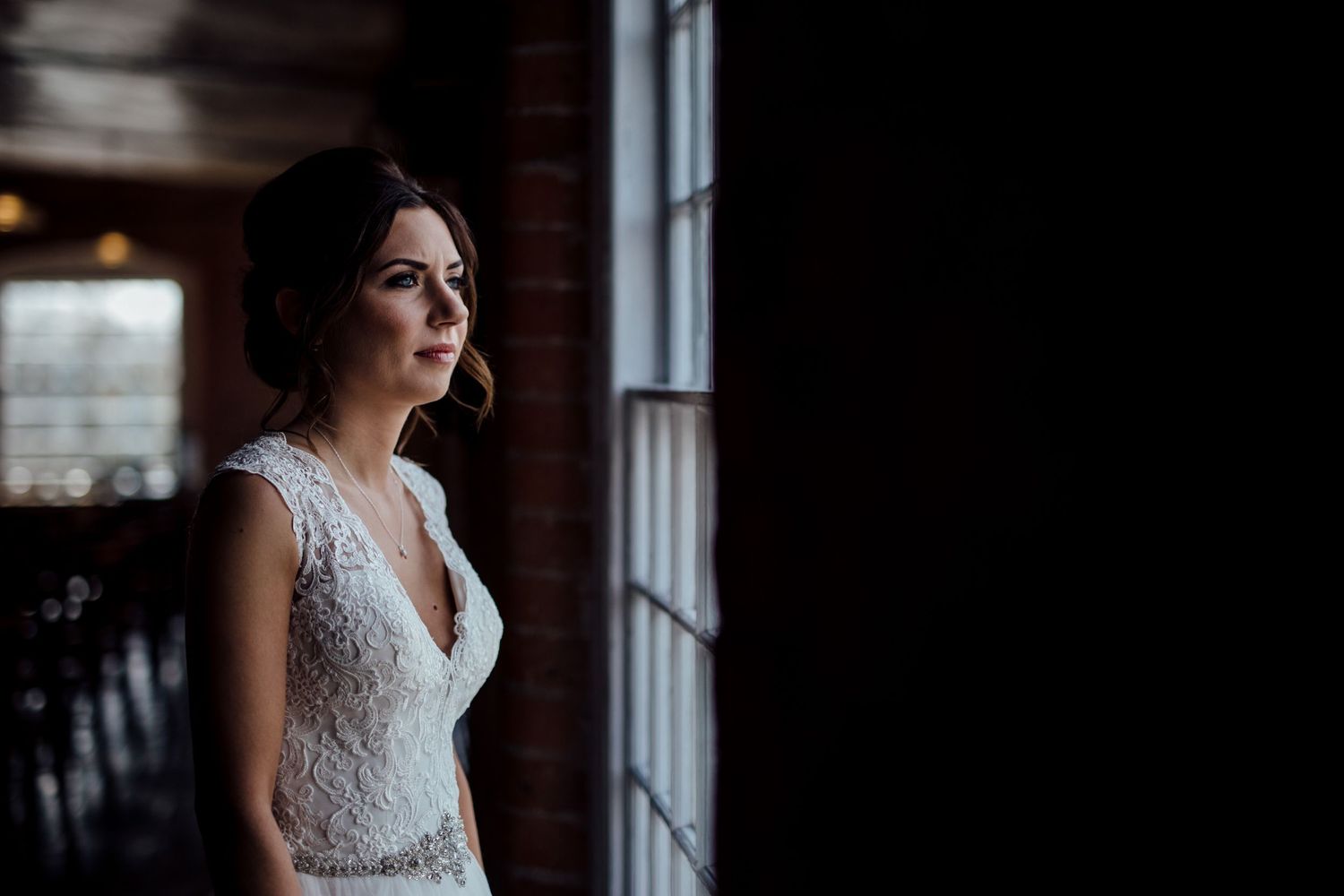 Bridal portrait in lace gown with dramatic window lighting and industrial brick backdrop.