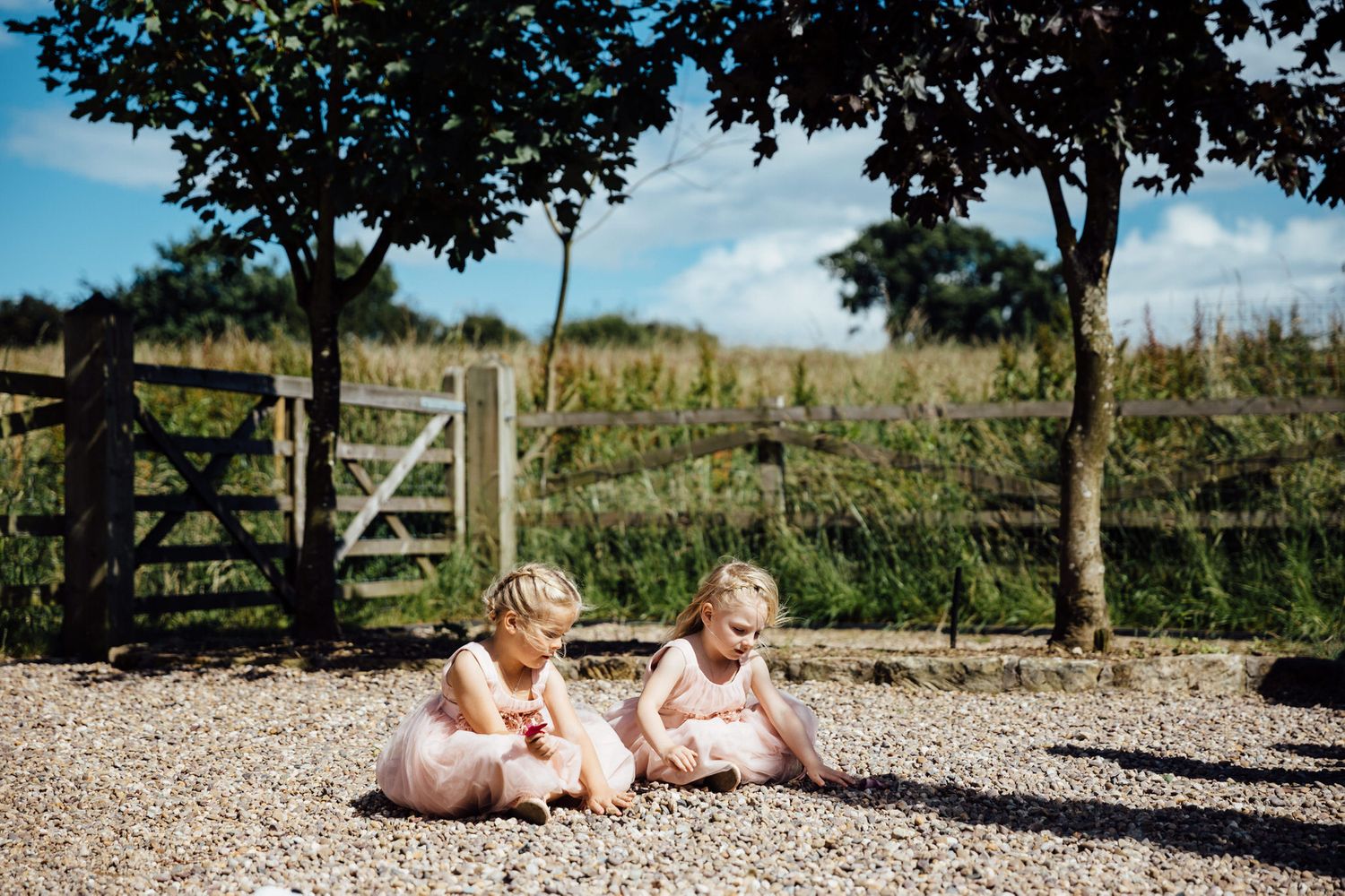 Two young children in pink dresses sitting on a gravel path near a wooden gate.