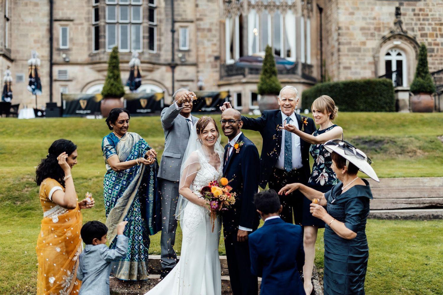 Wedding party and guests gather on lawn for group photos outside historic stone building.