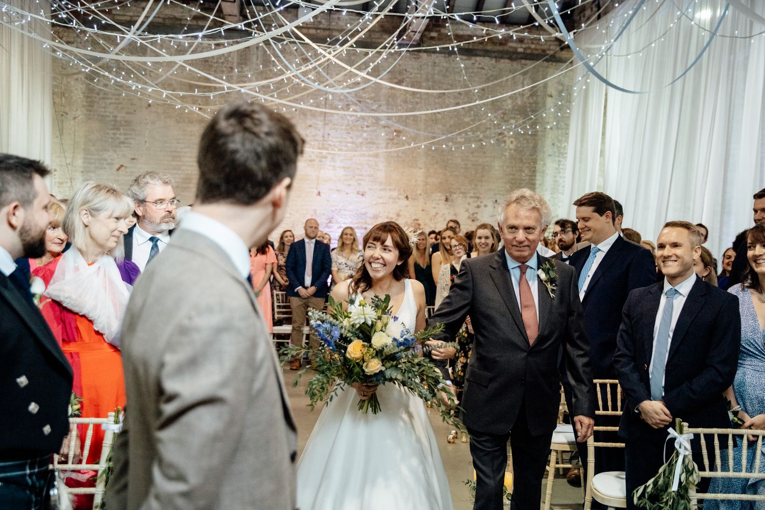 Wedding guests gather in an elegantly decorated venue with white drapes and string lights overhead.