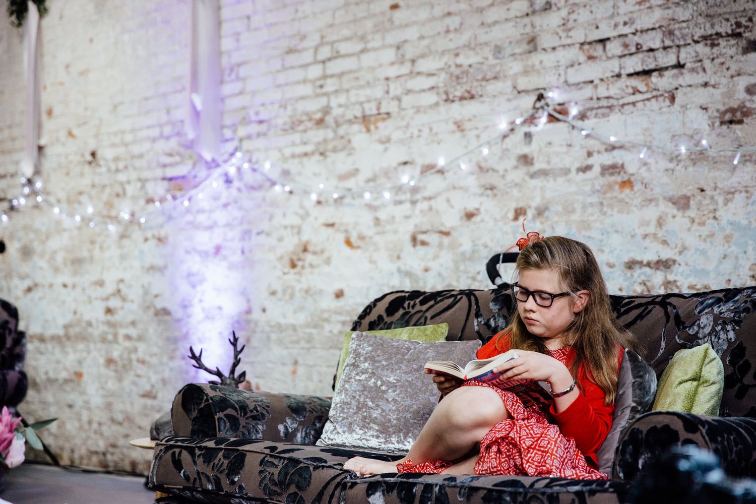 A person in red reads on a vintage couch against an exposed brick wall with purple lighting.
