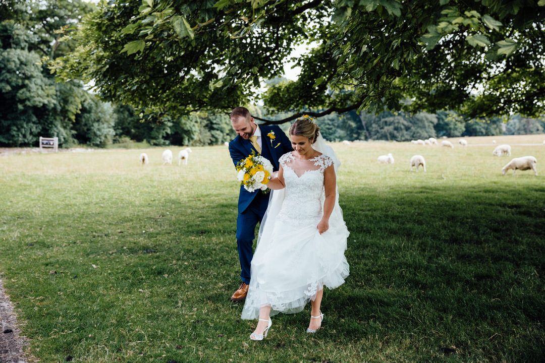 Wedding couple walks through lush green field with sheep grazing in background.