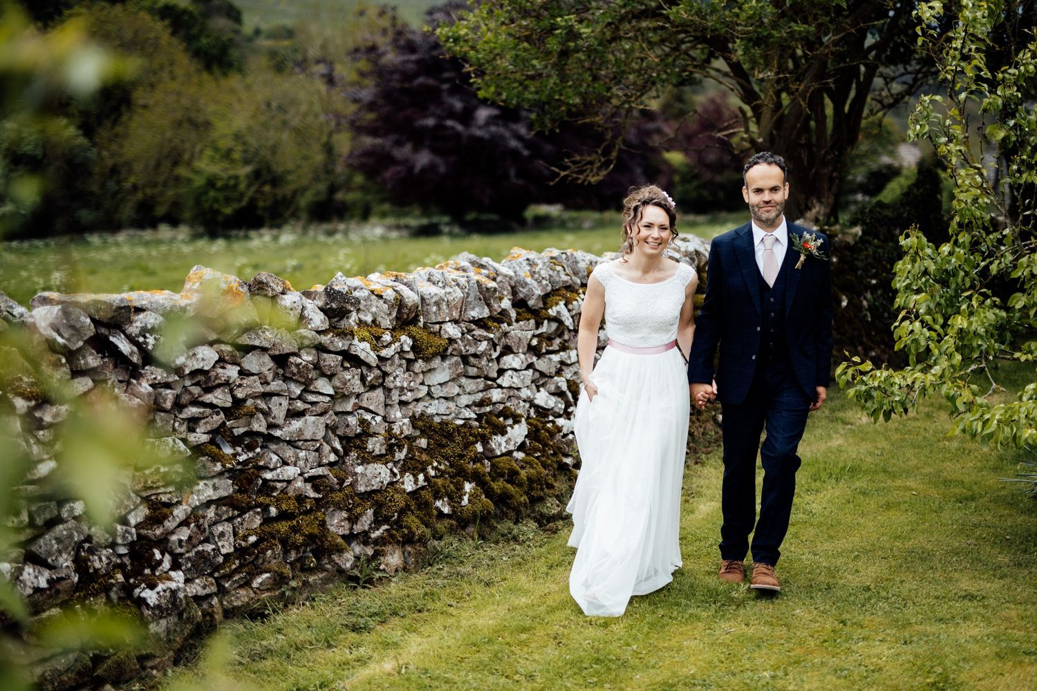 Couple walking through lush garden with stone wall backdrop.