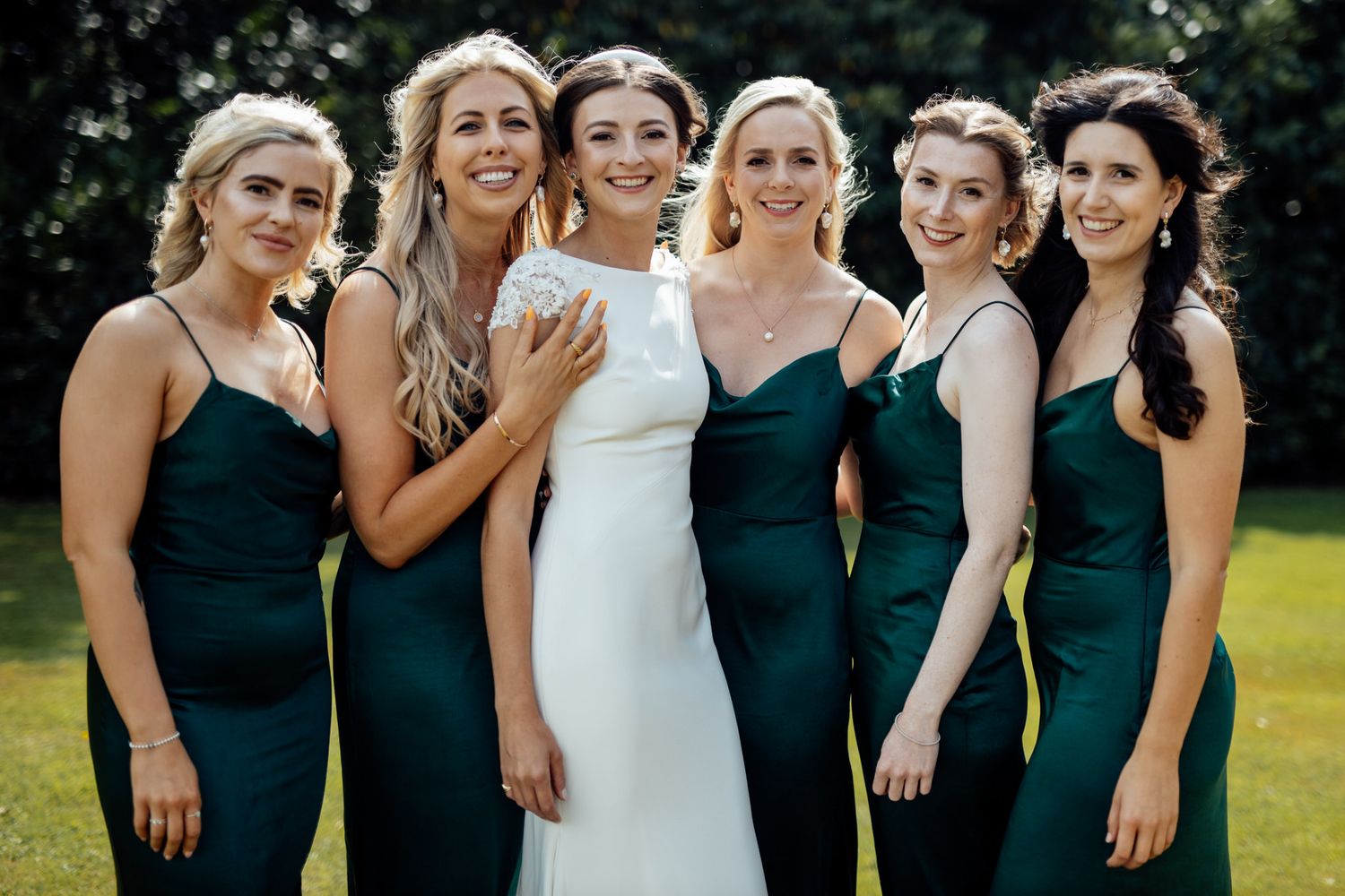 Bridesmaids in dark green dresses laughing together outdoors on a sunny day.